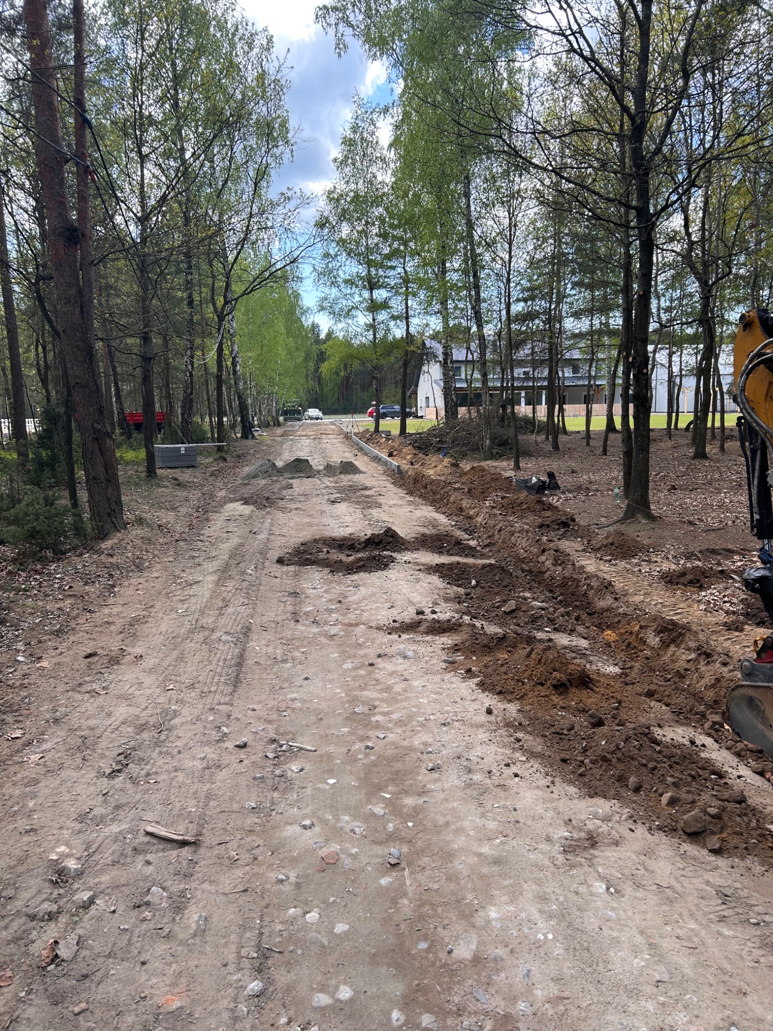 Wykop pod drogę leśną w trakcie realizacji, widoczny wykopany rów, przygotowane materiały budowlane i częściowo odsłonięte korzenie drzew.