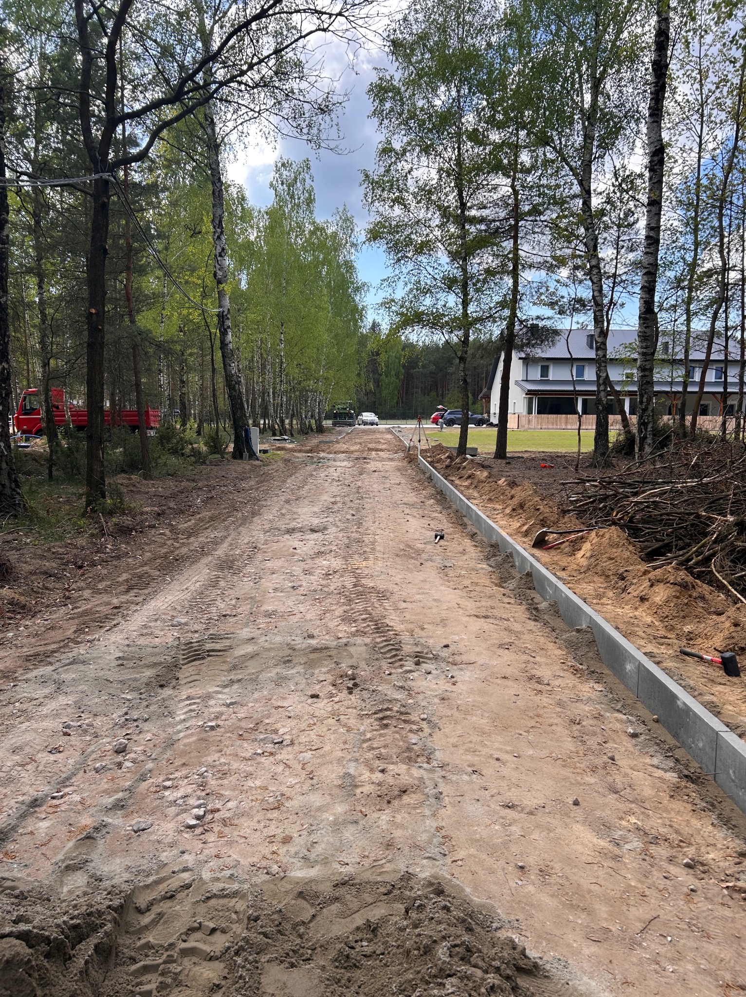 Układanie obrzeży chodnika wzdłuż drogi gruntowej na tle drzew i domu. Widoczne narzędzia i ślady opon. Prace drogowe w toku, etap budowy drogi.
