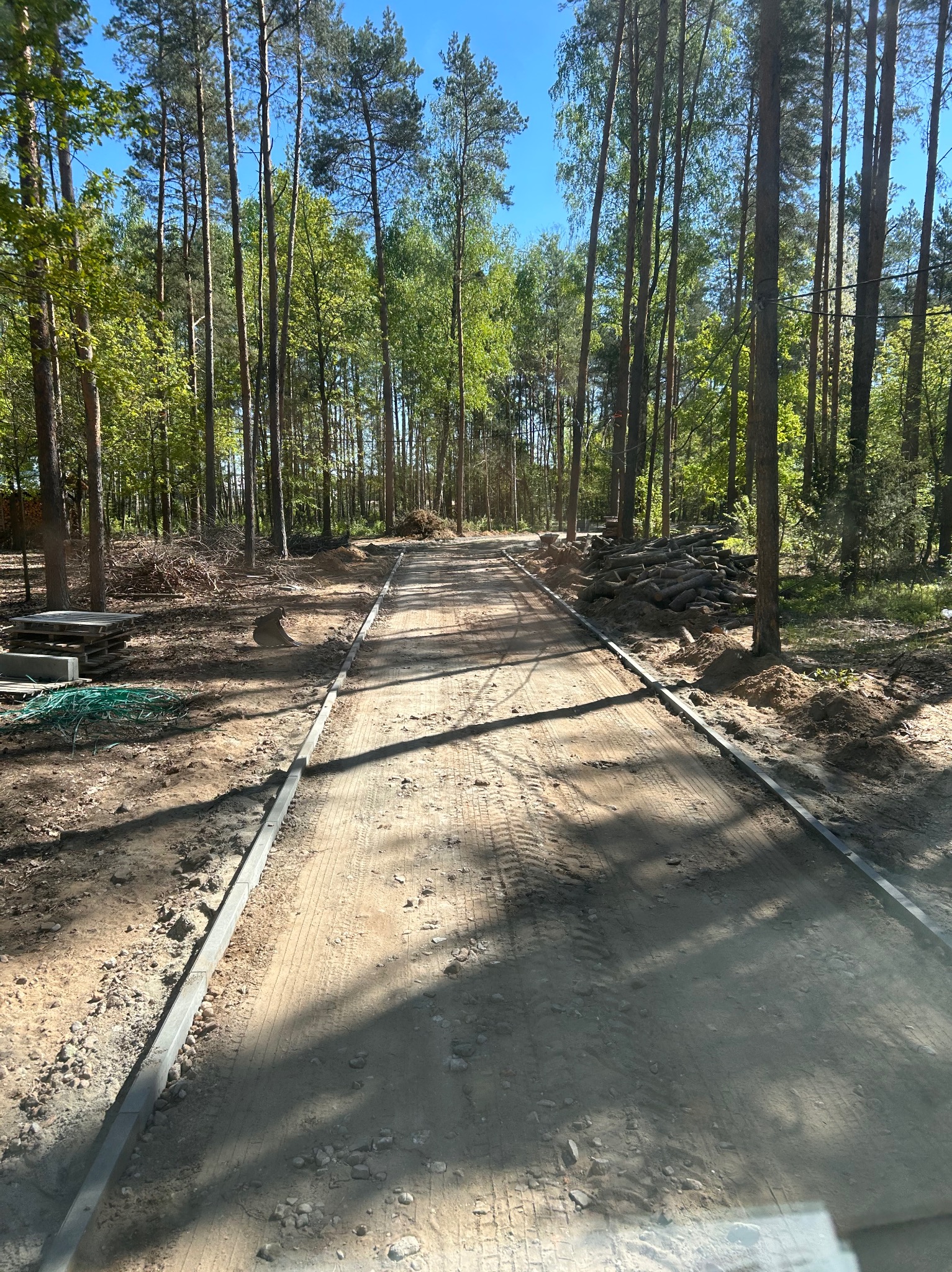Droga gruntowa w lesie z ułożonymi obrzeżami. Ślady opon na piasku, w tle stosy gałęzi i drzew, błękitne niebo. Prace ziemne w toku.