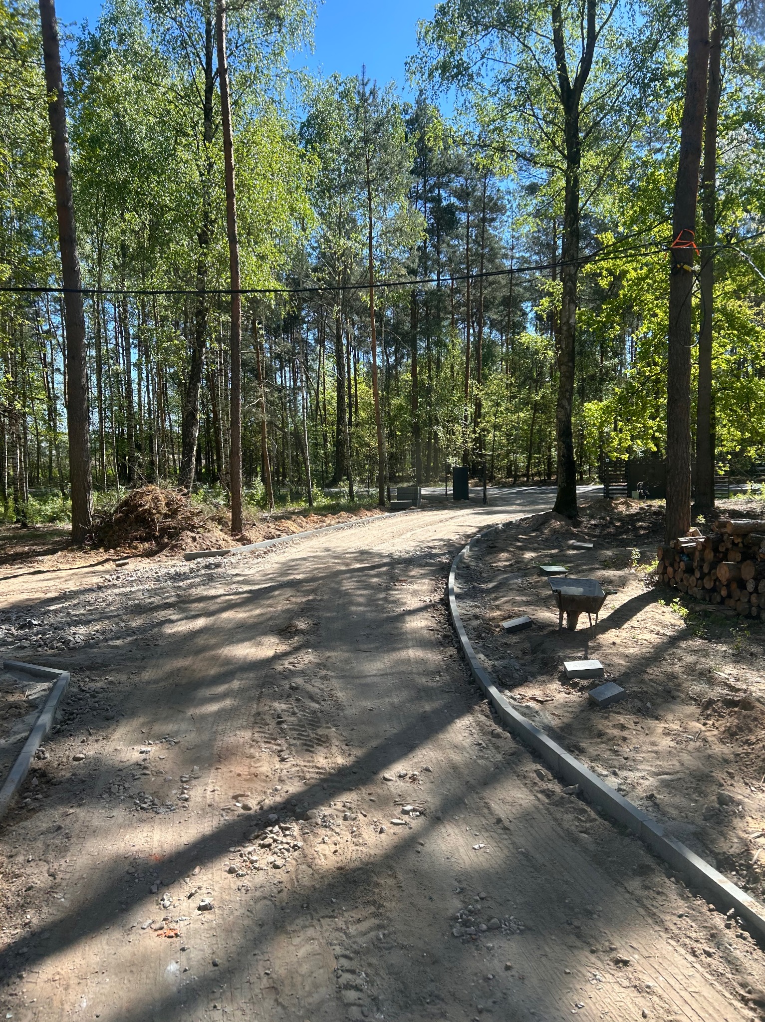 Układanie obrzeży drogi w Karniewo. Widok na gruntową drogę z ułożonym obrzeżem z kostki betonowej, w tle las i sterta gałęzi. Prace ziemne.