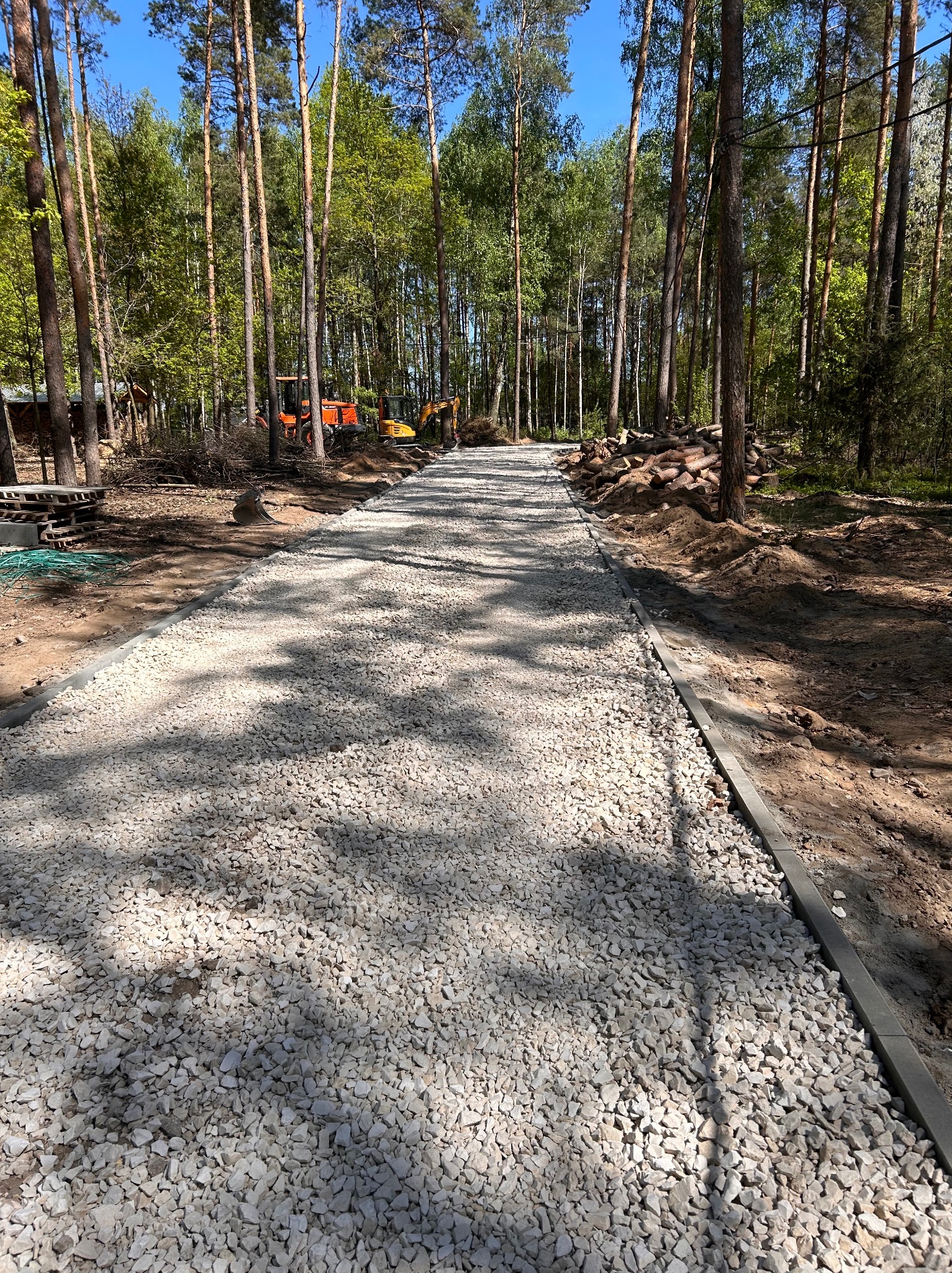 Utwardzona droga z białego kruszywa w lesie, obrzeża z szarego betonu. W tle maszyny budowlane i stosy drewna, wysokie drzewa.