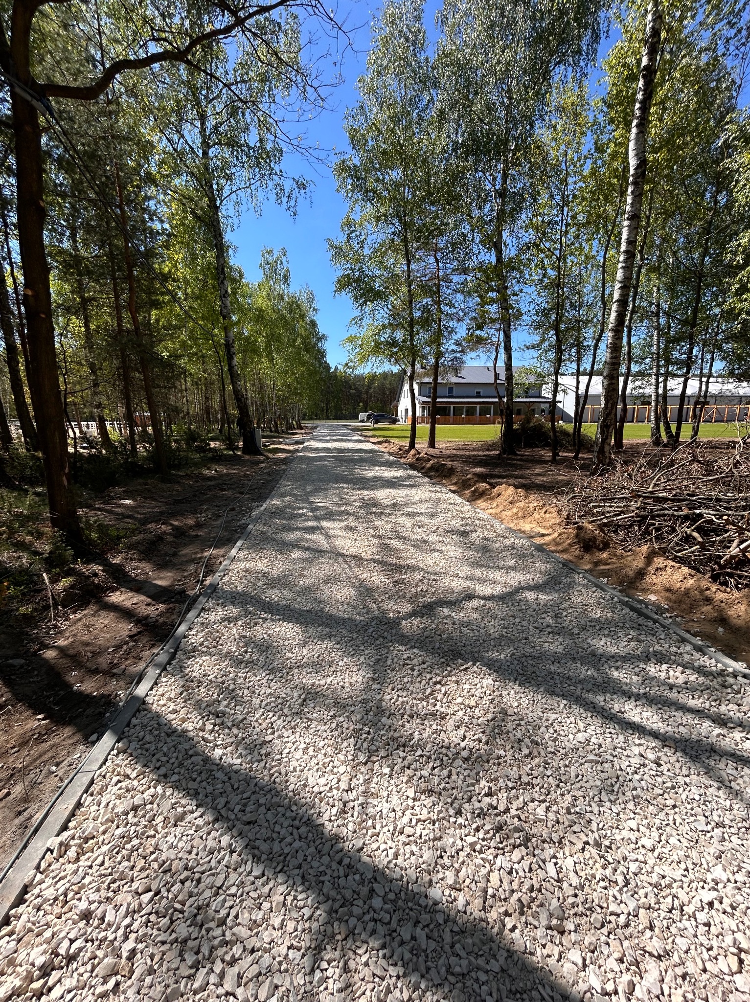 Utwardzona żwirowa droga wzdłuż drzew, zakończona obrzeżami, prowadząca do domu w oddali. Słoneczny dzień, wyraźne cienie drzew na żwirze.