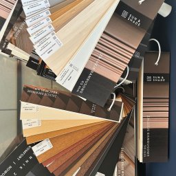 IDEAL ADRIANNA NOWACZYK - Paleta próbek żaluzji bambusowych i drewnianych marki Sun & Shade w różnych odcieniach i szerokościach, prezentowana na ciemnym tle.