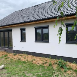 IDEAL ADRIANNA NOWACZYK - Nowoczesny dom jednorodzinny z grafitowymi oknami i roletami zewnętrznymi, widok z boku na elewację z widocznymi śladami prac ziemnych wokół budynku.