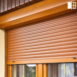 IDEAL ADRIANNA NOWACZYK - Brązowa roleta zewnętrzna częściowo zasłonięta, zamontowana na oknie z drewnianą ramą, widoczna na tle ceglanej ściany i logo producenta w prawym górnym rogu.