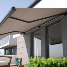 Beżowa markiza tarasowa z grafitową konstrukcją, zamontowana nad oknem w nowoczesnym domu. W tle biały budynek i błękitne niebo.