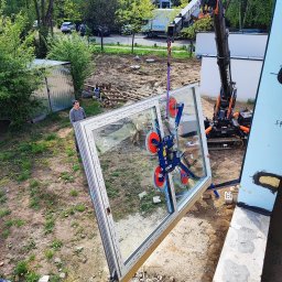 IDEAL ADRIANNA NOWACZYK - Montaż okna z użyciem dźwigu i przyssawki. Dwóch mężczyzn nadzoruje proces. Widoczne elementy konstrukcyjne budynku i otoczenie placu budowy.