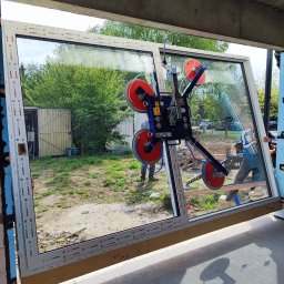 IDEAL ADRIANNA NOWACZYK - Montaż okna PCV z użyciem przyssawki próżniowej. Widoczni dwaj monterzy, okno z szybą refleksyjną i profilem Aluplast. Budowa w tle.