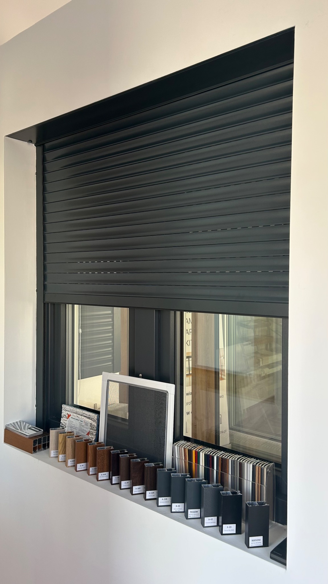 Zamknięta, grafitowa roleta zewnętrzna na oknie PCV z ekspozycją próbek kolorystycznych na parapecie, w tym drewnopodobnych i czarnych, wraz z ramką moskitiery.