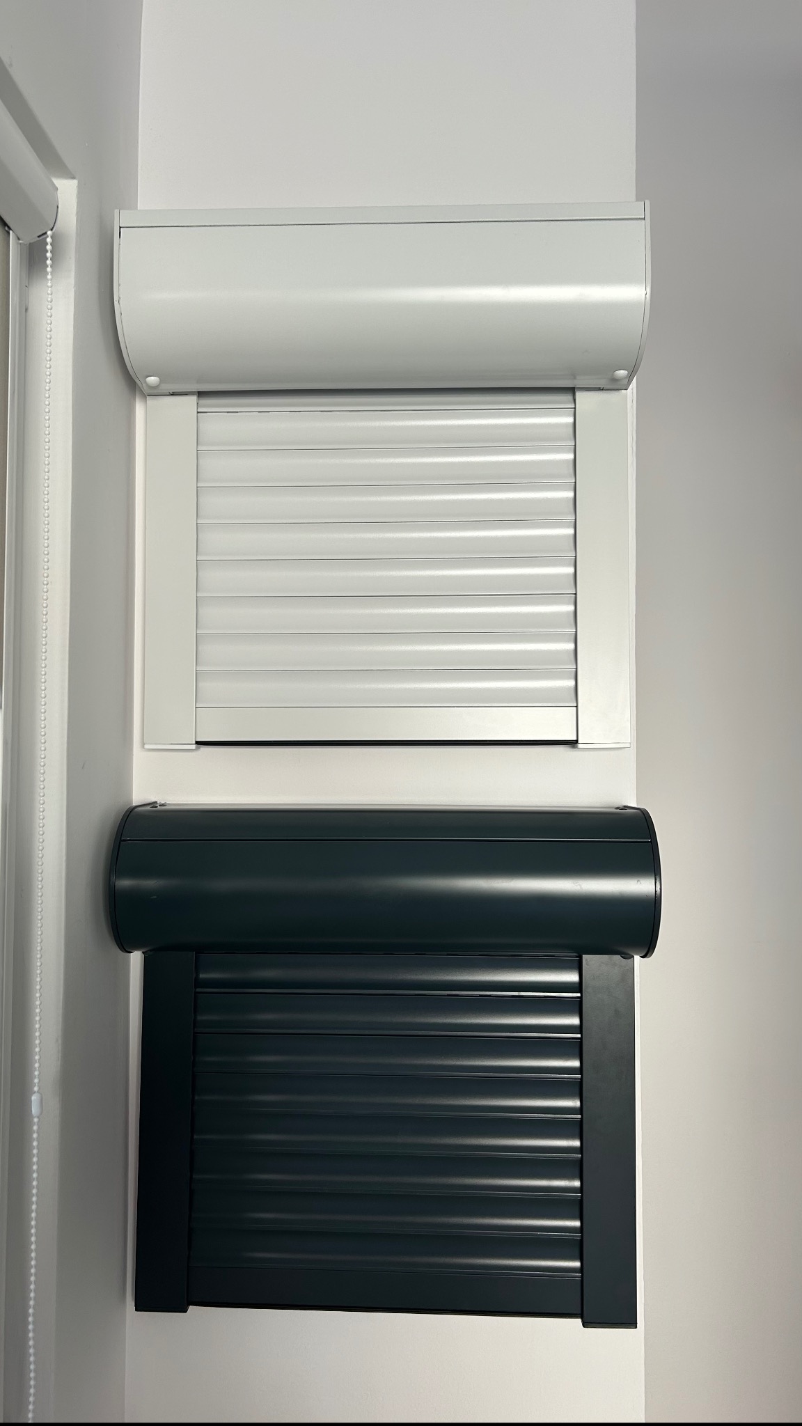 Dwie rolety zewnętrzne: biała i grafitowa, zamontowane jedna nad drugą na ścianie wewnątrz pomieszczenia, widoczne detale konstrukcyjne.