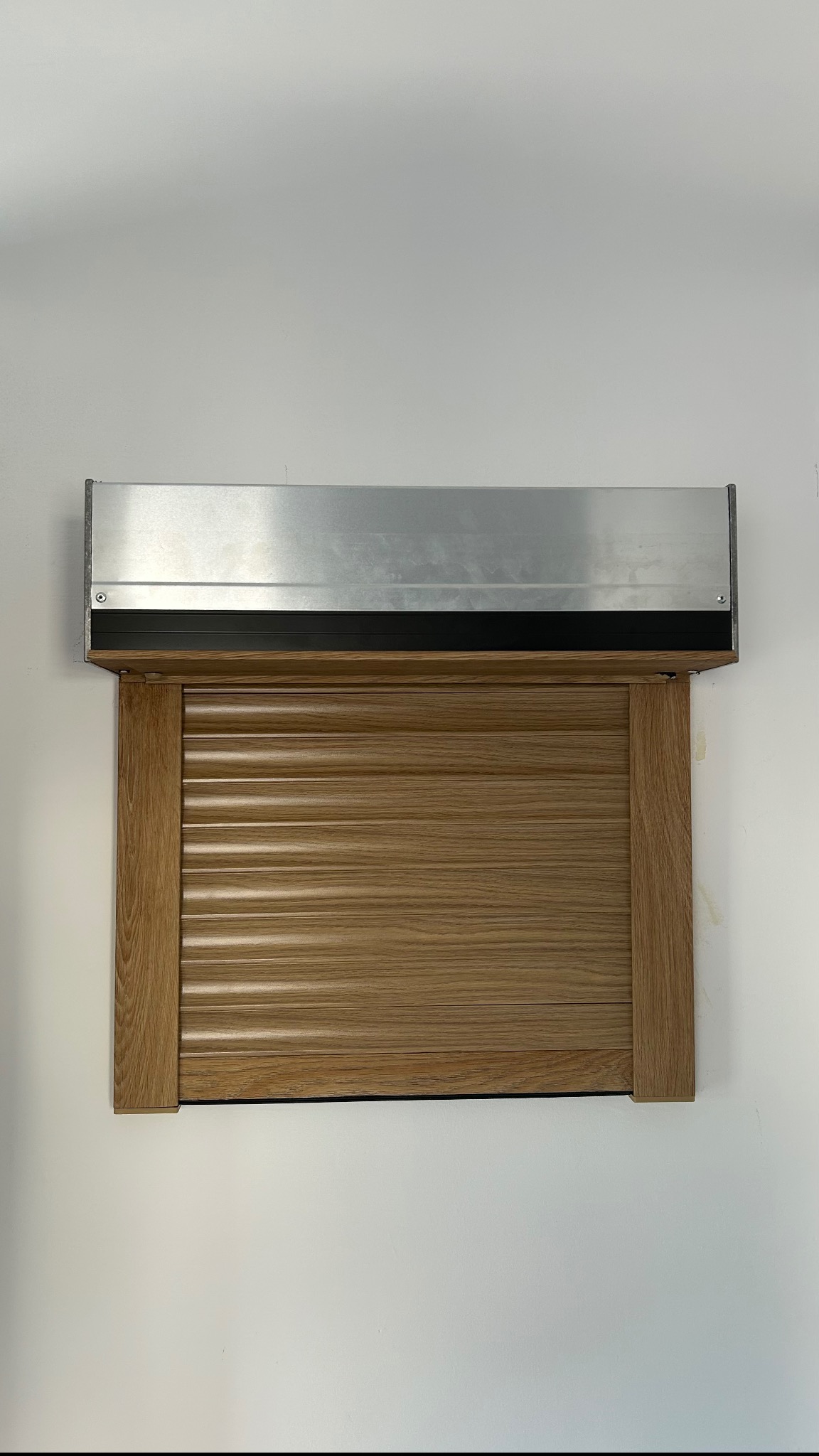 Zamknięta roleta zewnętrzna w kolorze jasnego drewna, zamontowana na białej ścianie, z widoczną aluminiową osłoną.