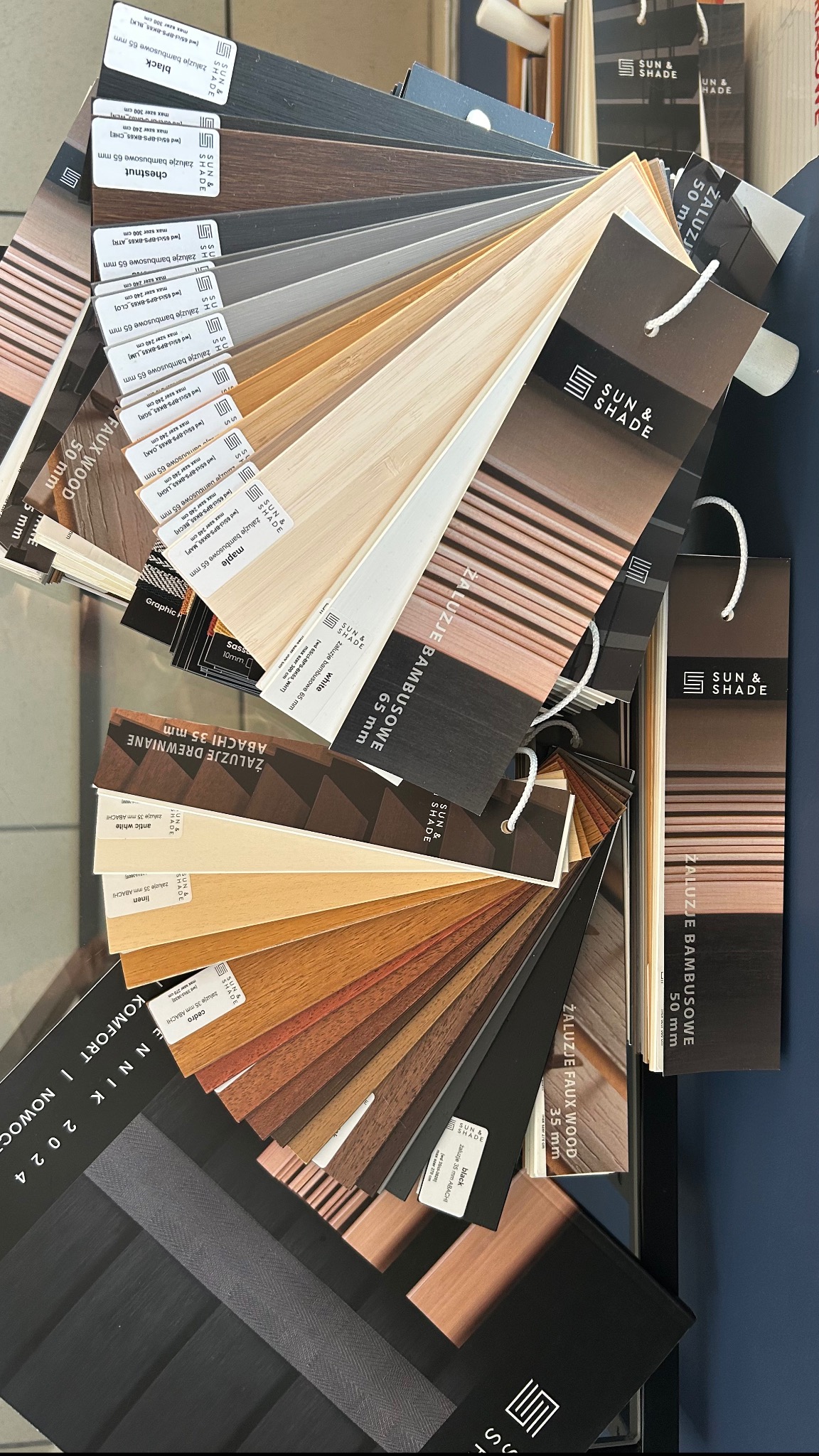 Paleta próbek żaluzji bambusowych i drewnianych marki Sun & Shade w różnych odcieniach i szerokościach, prezentowana na ciemnym tle.