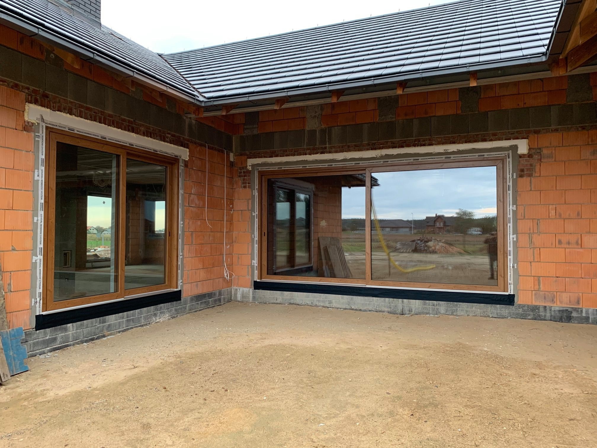 Montaż okien przesuwnych z brązowymi ramami w surowym stanie budynku z cegły, widoczne profile okienne Soudal.