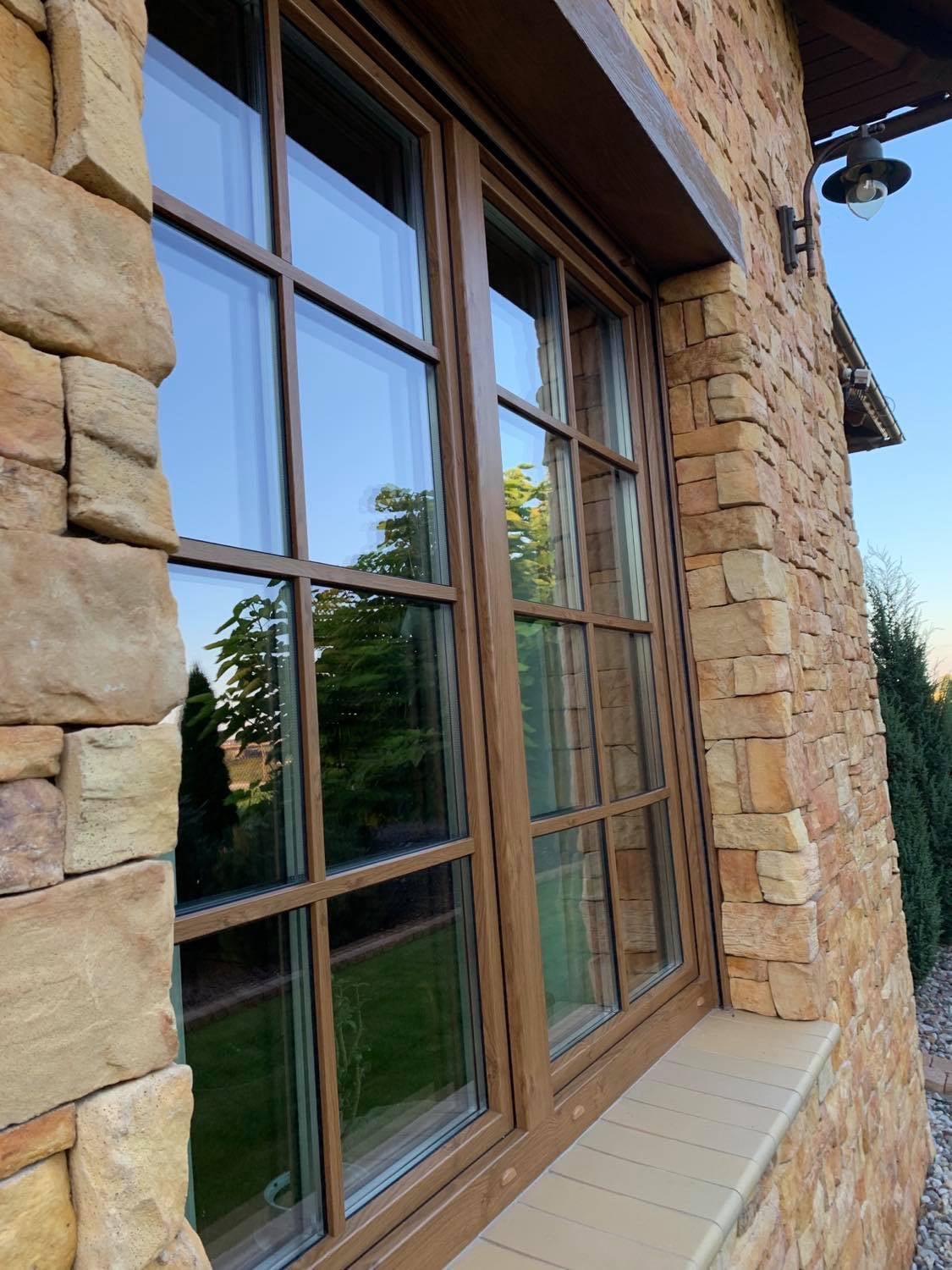 Brązowe okno z podziałami szprosowymi w elewacji z kamienia piaskowca, z widocznym odbiciem zieleni w szybach i fragmentem rolety zewnętrznej.