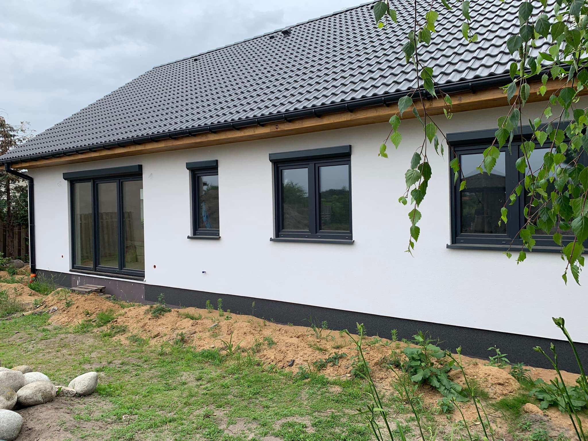 Nowoczesny dom jednorodzinny z grafitowymi oknami i roletami zewnętrznymi, widok z boku na elewację z widocznymi śladami prac ziemnych wokół budynku.