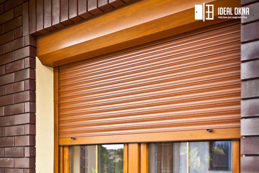 Brązowa roleta zewnętrzna częściowo zasłonięta, zamontowana na oknie z drewnianą ramą, widoczna na tle ceglanej ściany i logo producenta w prawym górnym rogu.