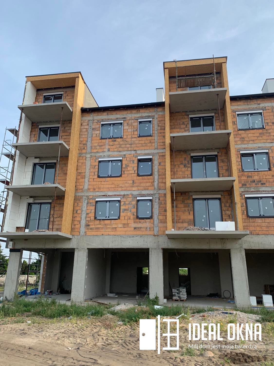 Budynek w trakcie budowy z zamontowanymi oknami o ciemnych ramach, widoczne rusztowania i materiały budowlane, na pierwszym planie logo firmy.