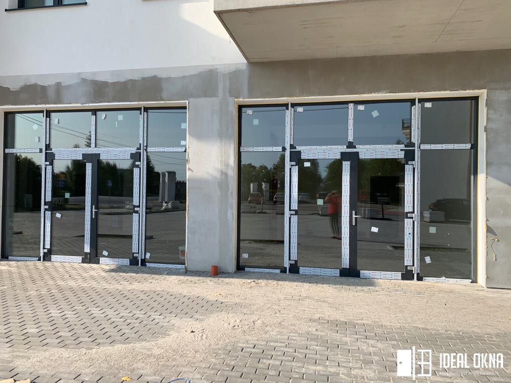 Dwa duże, przeszklone wejścia z czarnymi ramami, jeszcze zabezpieczone taśmą montażową, w surowym stanie budynku, kostka brukowa na pierwszym planie, logo firmy w prawym dolnym rogu.