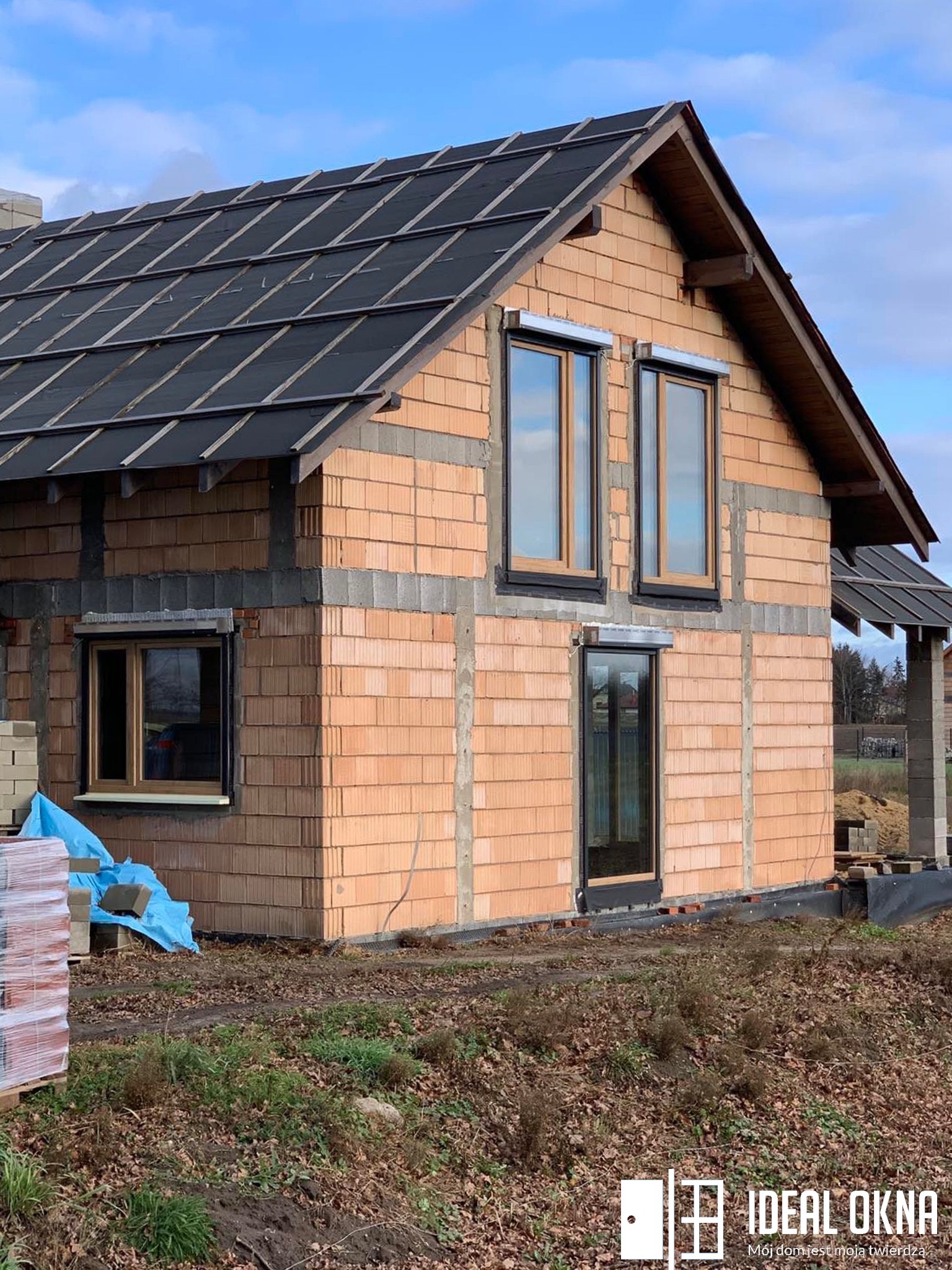 Surowy budynek w trakcie budowy z zamontowanymi oknami PCV i roletami zewnętrznymi, widoczny logo firmy Ideal Okna w prawym dolnym rogu.