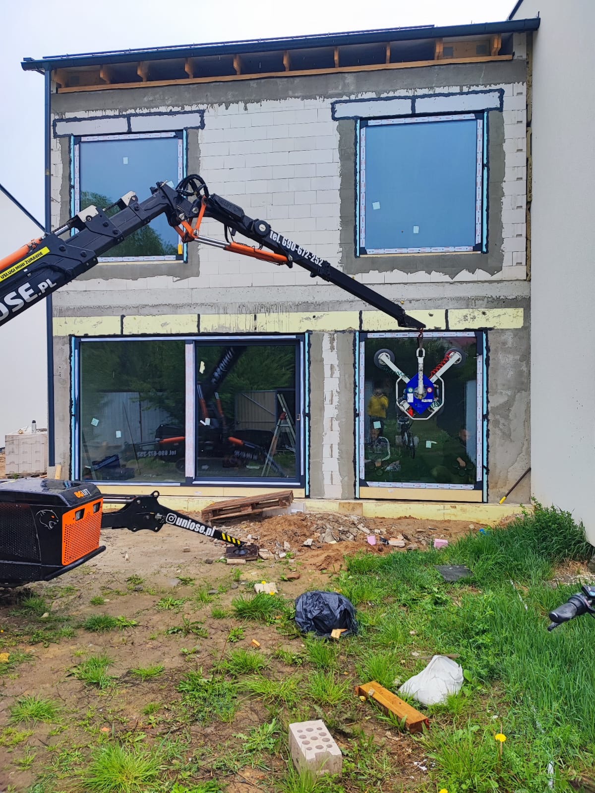 Montaż okna z użyciem dźwigu przyssawkowego na budowie domu. Widoczne okna na różnych etapach instalacji, ściany z betonu komórkowego.