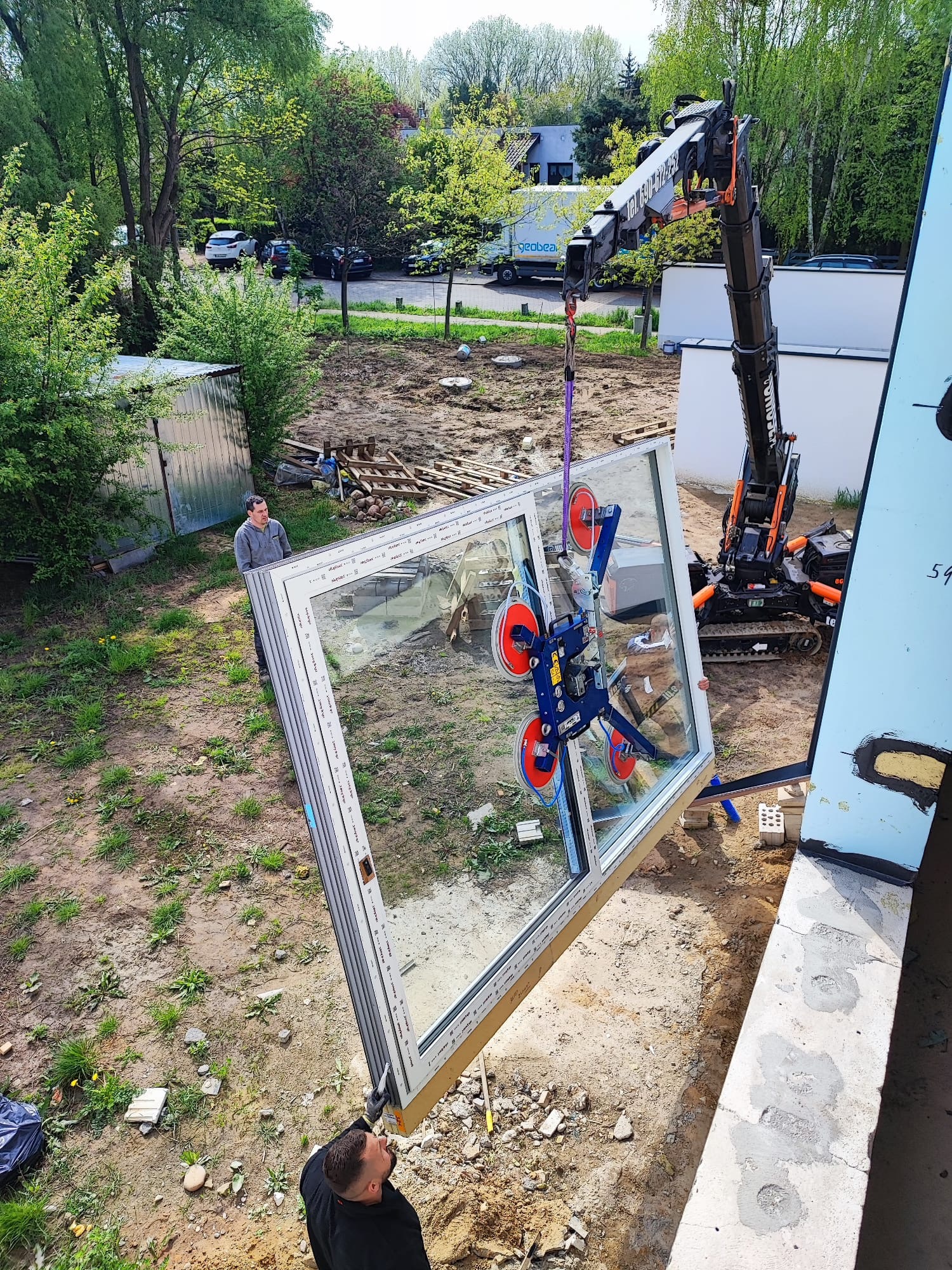 Montaż okna z użyciem dźwigu i przyssawki. Dwóch mężczyzn nadzoruje proces. Widoczne elementy konstrukcyjne budynku i otoczenie placu budowy.