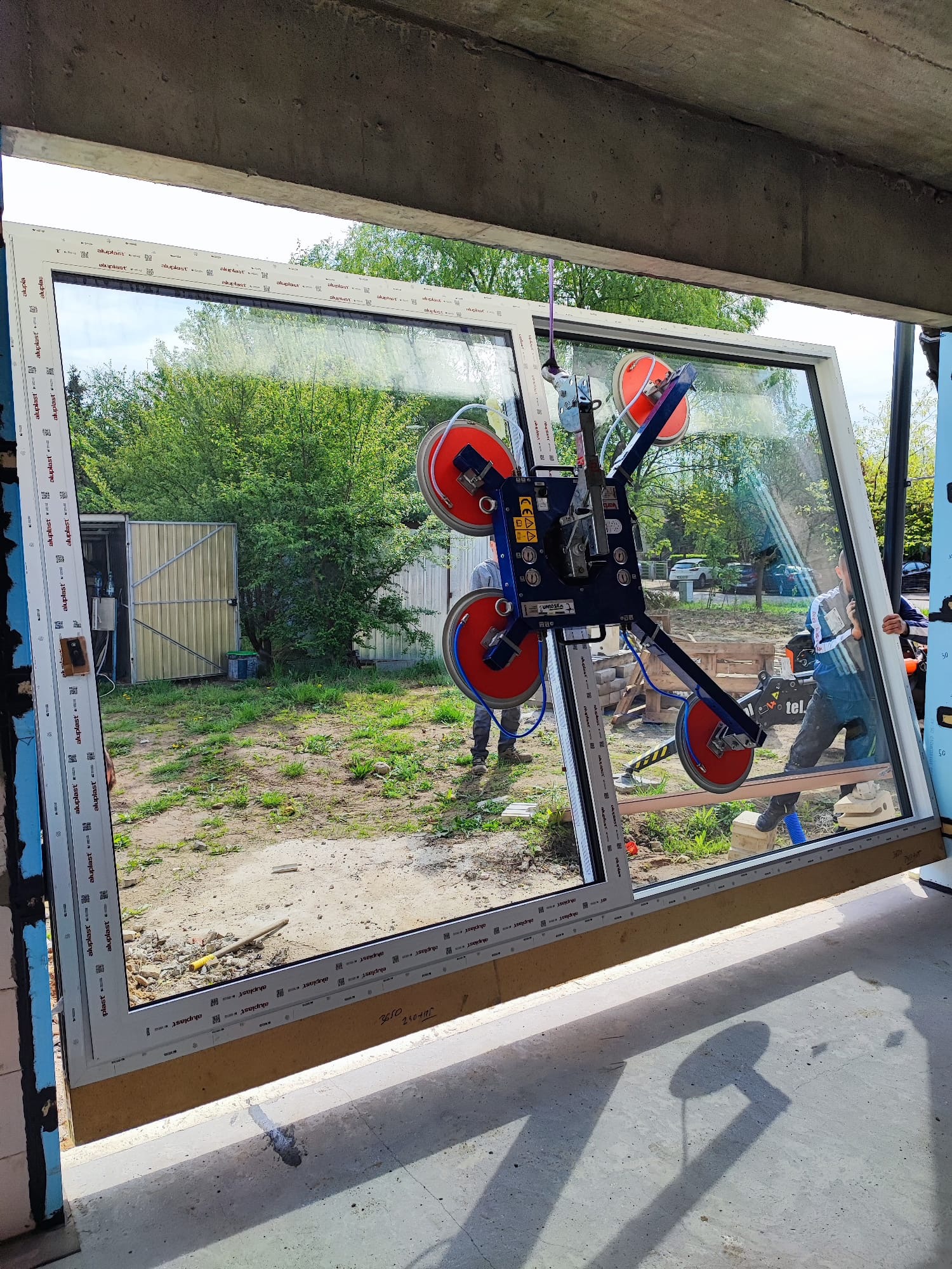 Montaż okna PCV z użyciem przyssawki próżniowej. Widoczni dwaj monterzy, okno z szybą refleksyjną i profilem Aluplast. Budowa w tle.