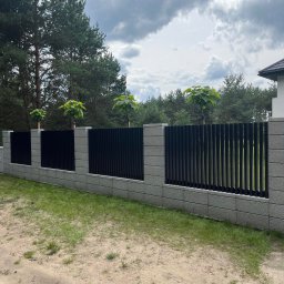 KULESZA WELD Kamil Kulesza - Nowoczesne ogrodzenie panelowe z czarnymi pionowymi panelami i szarymi słupkami z bloczków betonowych, z młodymi drzewkami posadzonymi wzdłuż ogrodzenia, na tle zieleni i pochmurnego nieba.