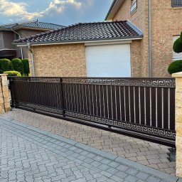 KULESZA WELD Kamil Kulesza - Przesuwna brama wjazdowa z metalowymi sztachetami i dekoracyjnym wzorem greckim, zamontowana przed ceglanym domem z białą bramą garażową.