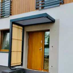 Nowoczesne wejście do domu z drewnianymi żaluzjami, zadaszeniem z metalową kratownicą i drzwiami w odcieniu miodowego dębu, widoczne wystające kable instalacji elektrycznej na elewacji.