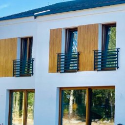 KULESZA WELD Kamil Kulesza - Nowoczesny dom z białymi ścianami, drewnianymi panelami i czarnymi, minimalistycznymi balustradami balkonowymi, widok na plac budowy.