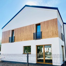 KULESZA WELD Kamil Kulesza - Nowoczesny dom z elewacją w kolorze białym i drewnopodobnym, z czarnymi balustradami balkonowymi i ogrodzeniem panelowym w trakcie montażu.