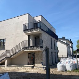 KULESZA WELD Kamil Kulesza - Nowoczesny budynek z balkonami i zewnętrznymi schodami z czarną metalową balustradą, worki z materiałami budowlanymi na placu budowy, błękitne niebo w tle.