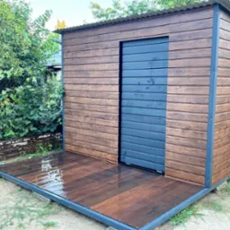 Nowoczesna, drewniana wiata ogrodowa z przesuwanymi, grafitowymi drzwiami i podestem, otoczona zielenią i elementami ogrodzenia z siatki.