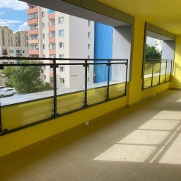 KULESZA WELD Kamil Kulesza - Żółty balkon z nowoczesną szklaną balustradą z czarnymi elementami mocującymi, widok na budynki mieszkalne i drzewa.
