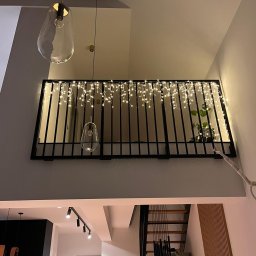 KULESZA WELD Kamil Kulesza - Wewnętrzna balustrada z czarnych metalowych prętów, ozdobiona girlandą świetlną, widok z dołu na antresolę z rośliną w tle, oświetlona wiszącymi lampami o organicznym kształcie.