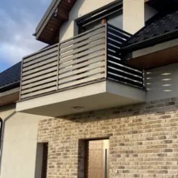 Nowoczesny balkon z poziomą balustradą z metalowych profili na tle elewacji z cegły klinkierowej i jasnego tynku.