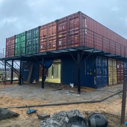 KULESZA WELD Kamil Kulesza - Dwukondygnacyjny budynek z kontenerów morskich w trakcie budowy, z metalową konstrukcją balkonu i schodami, otoczony piaskiem i materiałami budowlanymi.