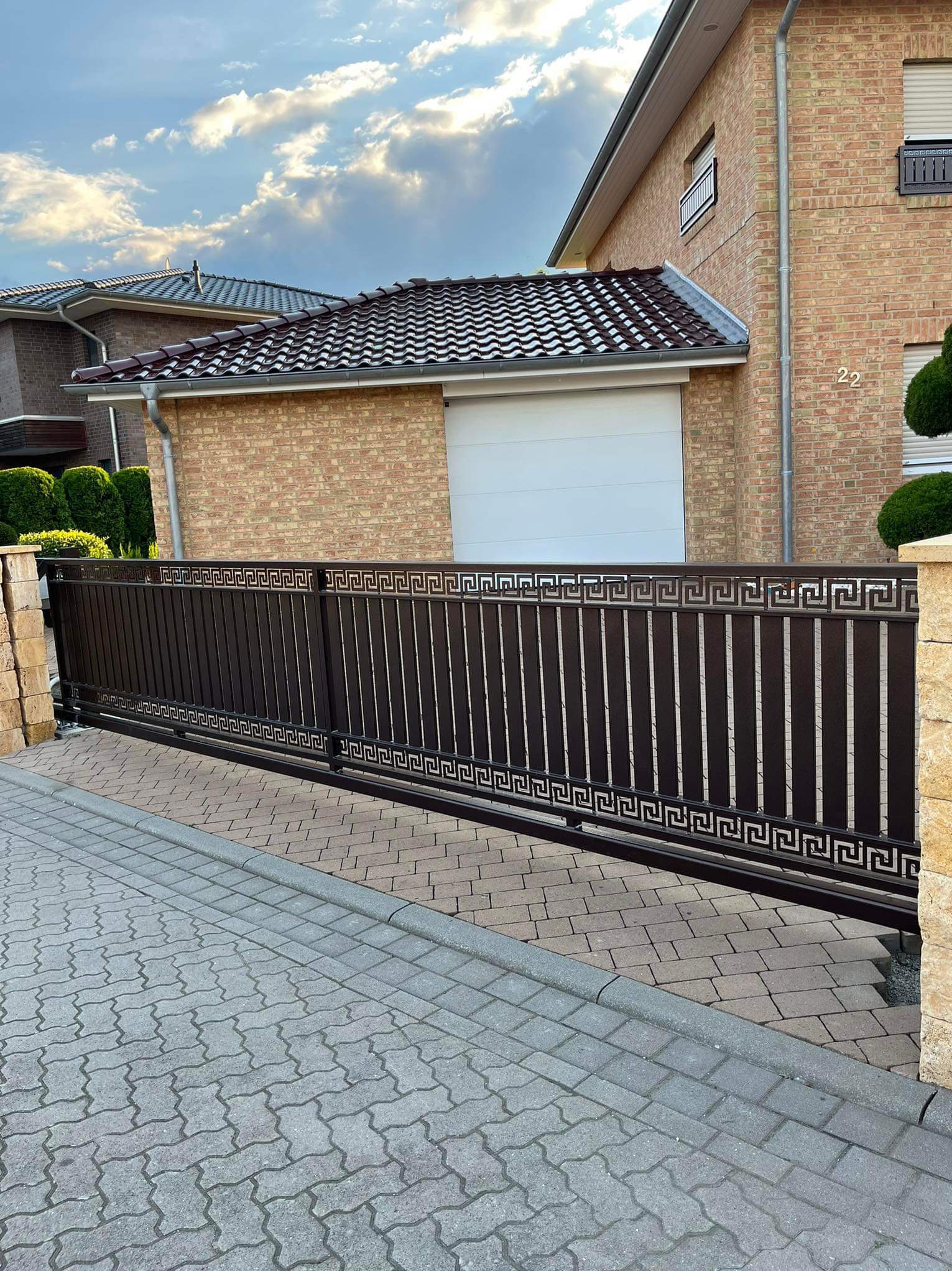 Przesuwna brama wjazdowa z metalowymi sztachetami i dekoracyjnym wzorem greckim, zamontowana przed ceglanym domem z białą bramą garażową.