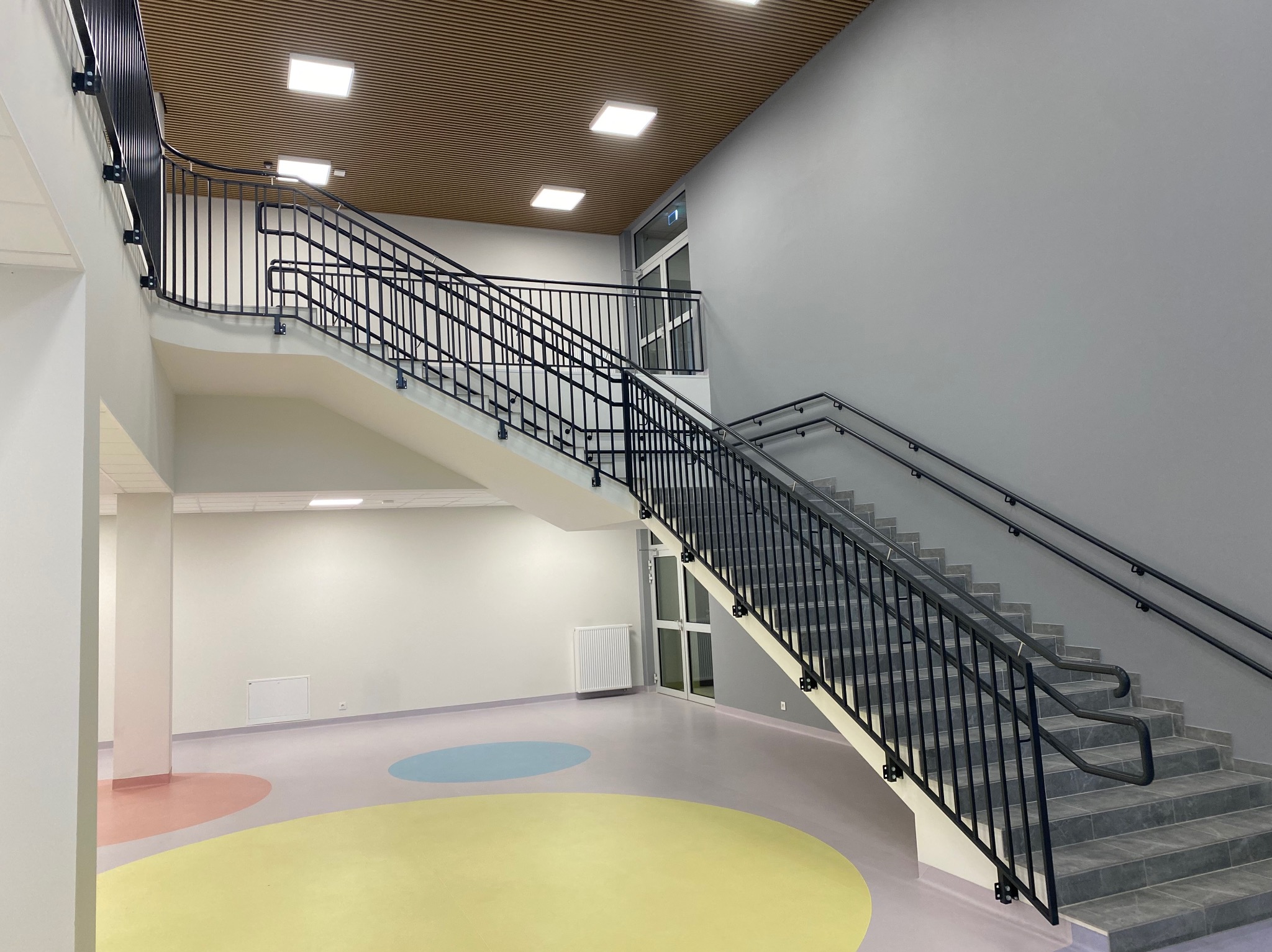 Wnętrze budynku z dwubiegowymi schodami o stalowej konstrukcji i czarnych, metalowych balustradach, widoczne kolorowe okręgi na podłodze i drewniany sufit.