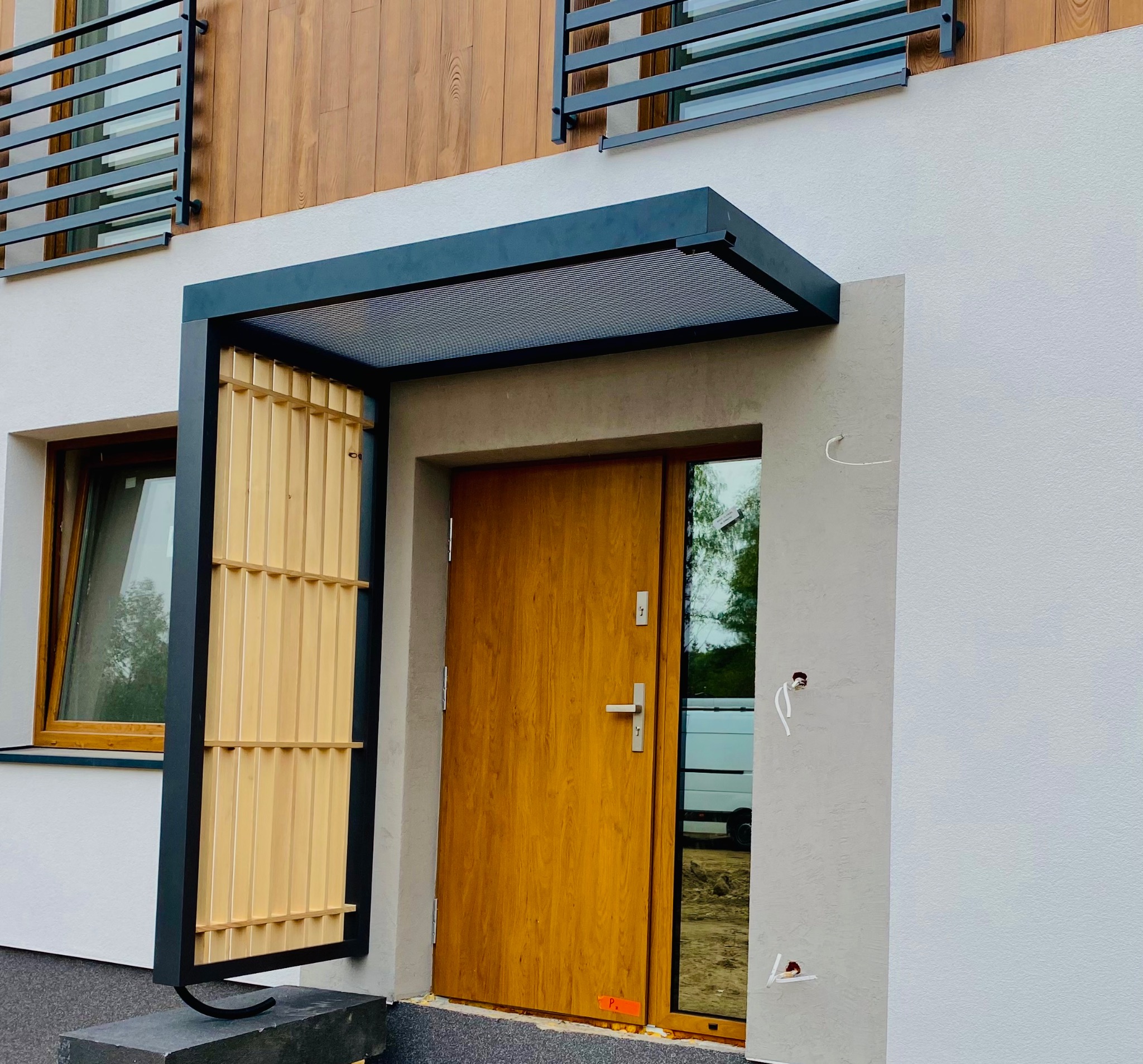 Nowoczesne wejście do domu z drewnianymi żaluzjami, zadaszeniem z metalową kratownicą i drzwiami w odcieniu miodowego dębu, widoczne wystające kable instalacji elektrycznej na elewacji.
