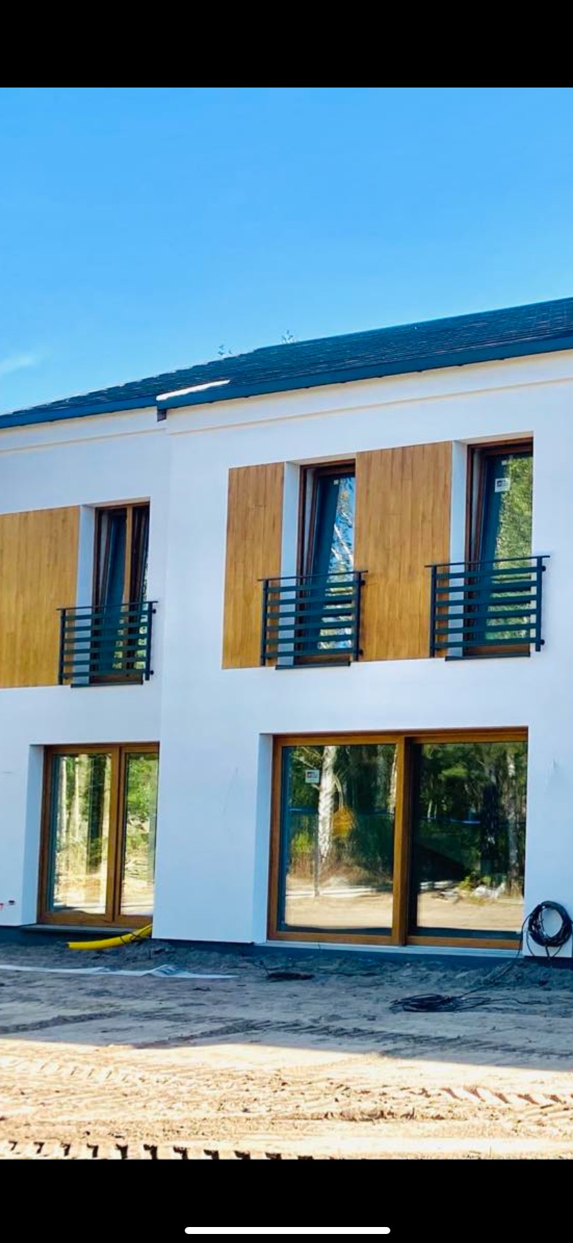 Nowoczesny dom z białymi ścianami, drewnianymi panelami i czarnymi, minimalistycznymi balustradami balkonowymi, widok na plac budowy.