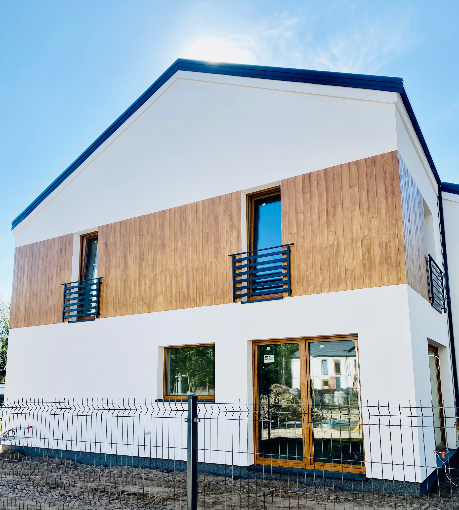 Nowoczesny dom z elewacją w kolorze białym i drewnopodobnym, z czarnymi balustradami balkonowymi i ogrodzeniem panelowym w trakcie montażu.