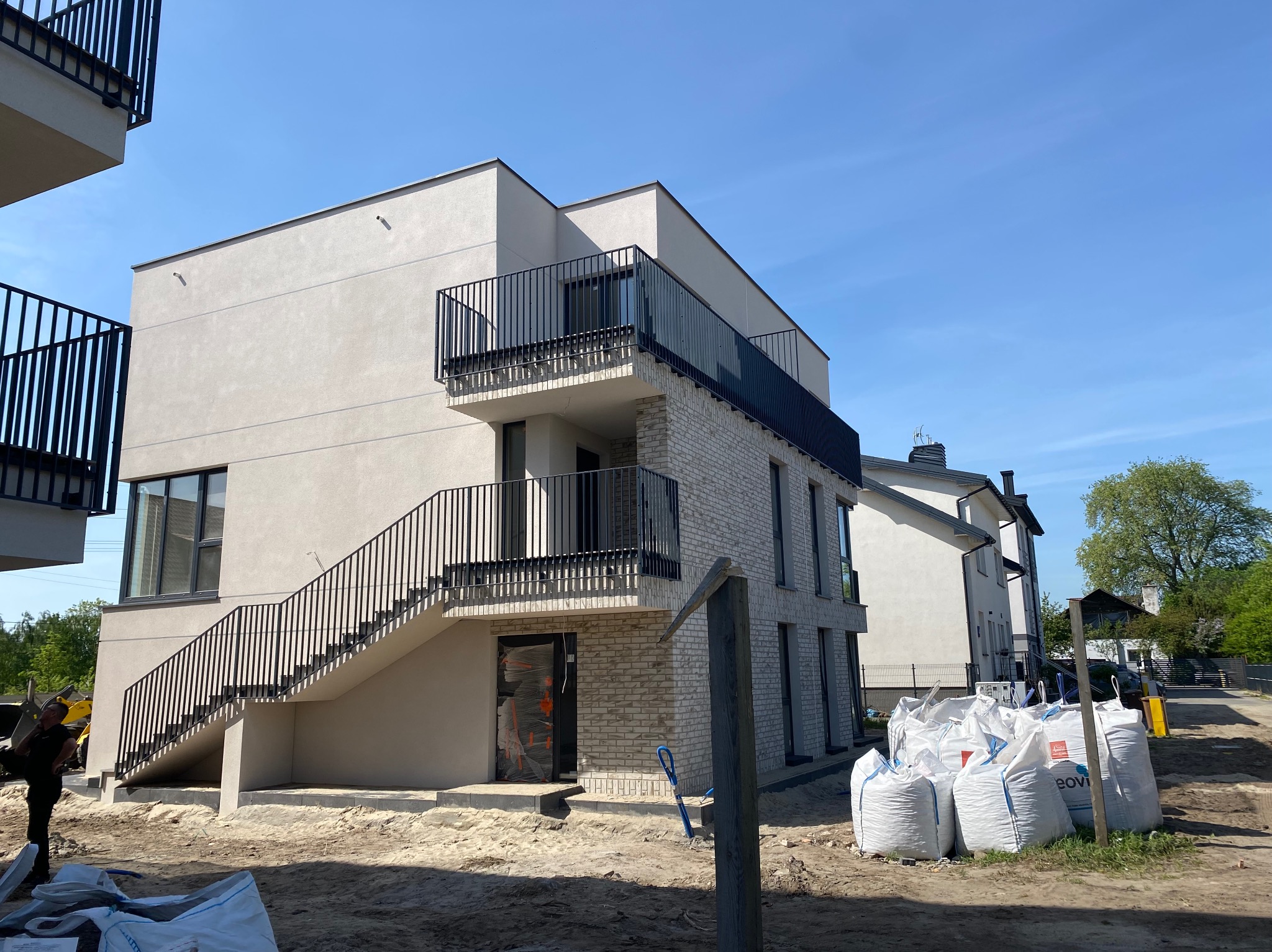 Nowoczesny budynek z balkonami i zewnętrznymi schodami z czarną metalową balustradą, worki z materiałami budowlanymi na placu budowy, błękitne niebo w tle.
