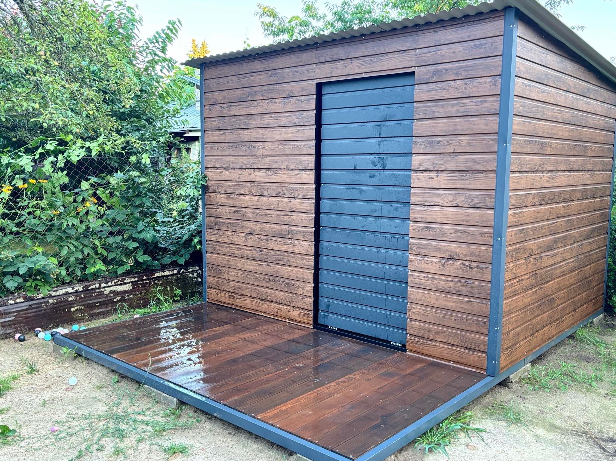 Nowoczesna, drewniana wiata ogrodowa z przesuwanymi, grafitowymi drzwiami i podestem, otoczona zielenią i elementami ogrodzenia z siatki.