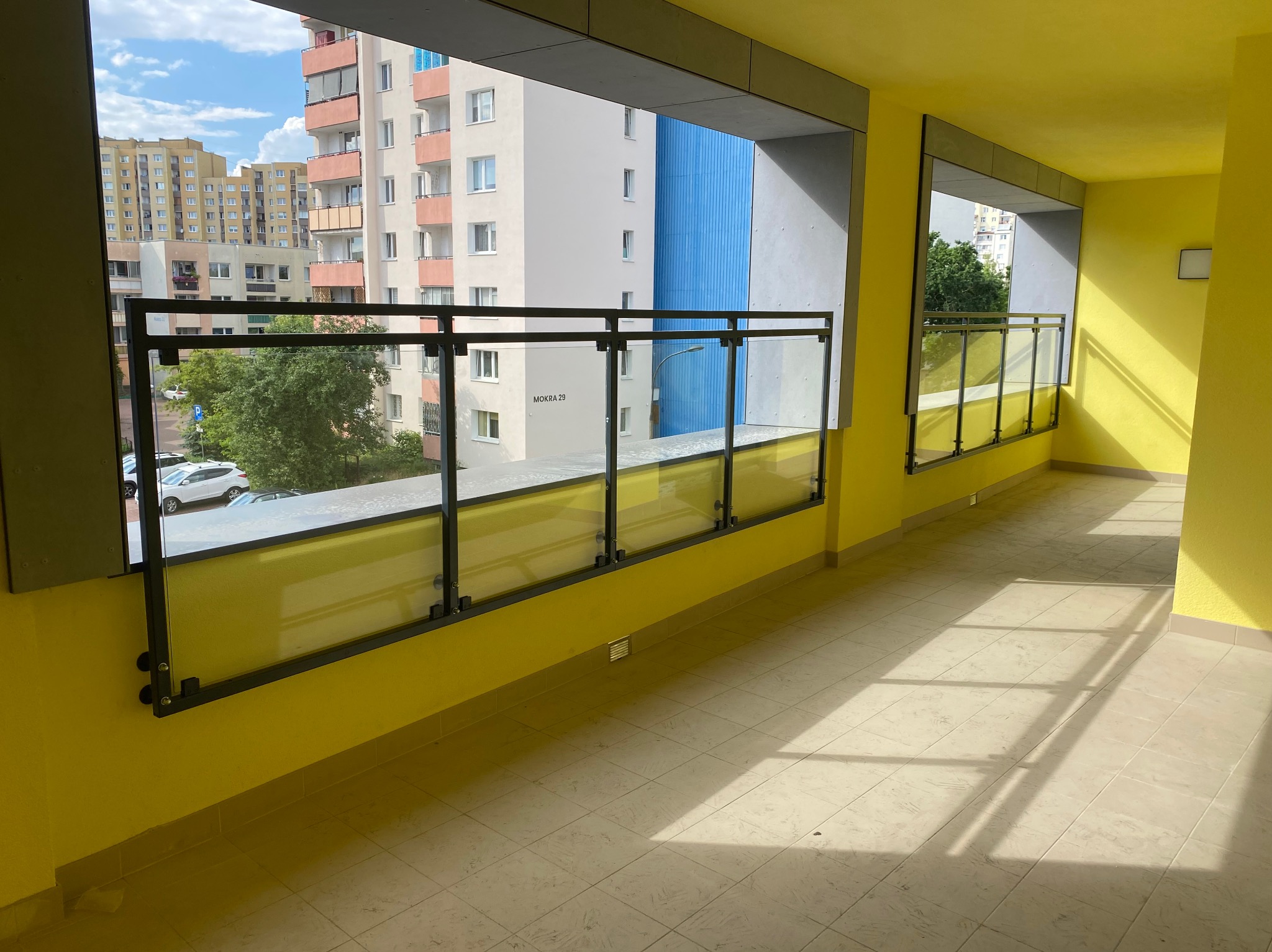 Żółty balkon z nowoczesną szklaną balustradą z czarnymi elementami mocującymi, widok na budynki mieszkalne i drzewa.