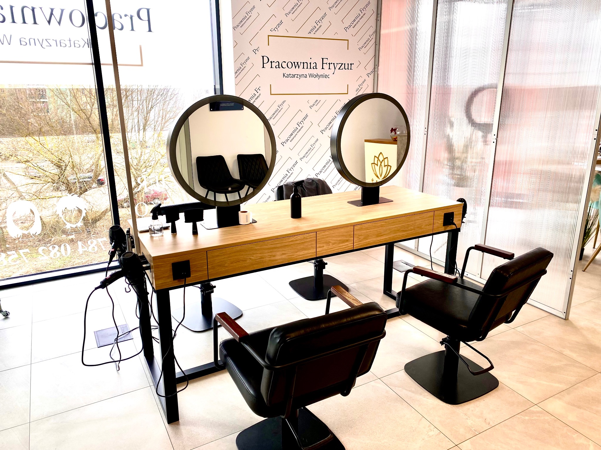 Wnętrze salonu fryzjerskiego: stanowiska z okrągłymi lustrami, drewnianymi blatami i czarnymi fotelami na tle ściany z logo firmy.