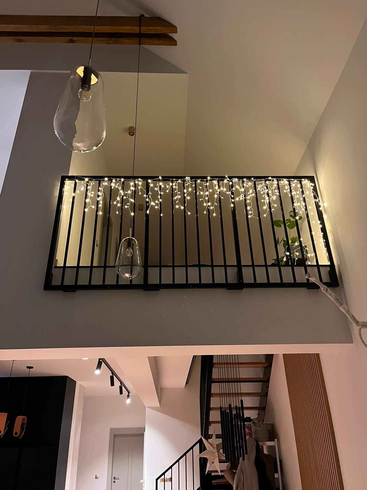 Wewnętrzna balustrada z czarnych metalowych prętów, ozdobiona girlandą świetlną, widok z dołu na antresolę z rośliną w tle, oświetlona wiszącymi lampami o organicznym kształcie.