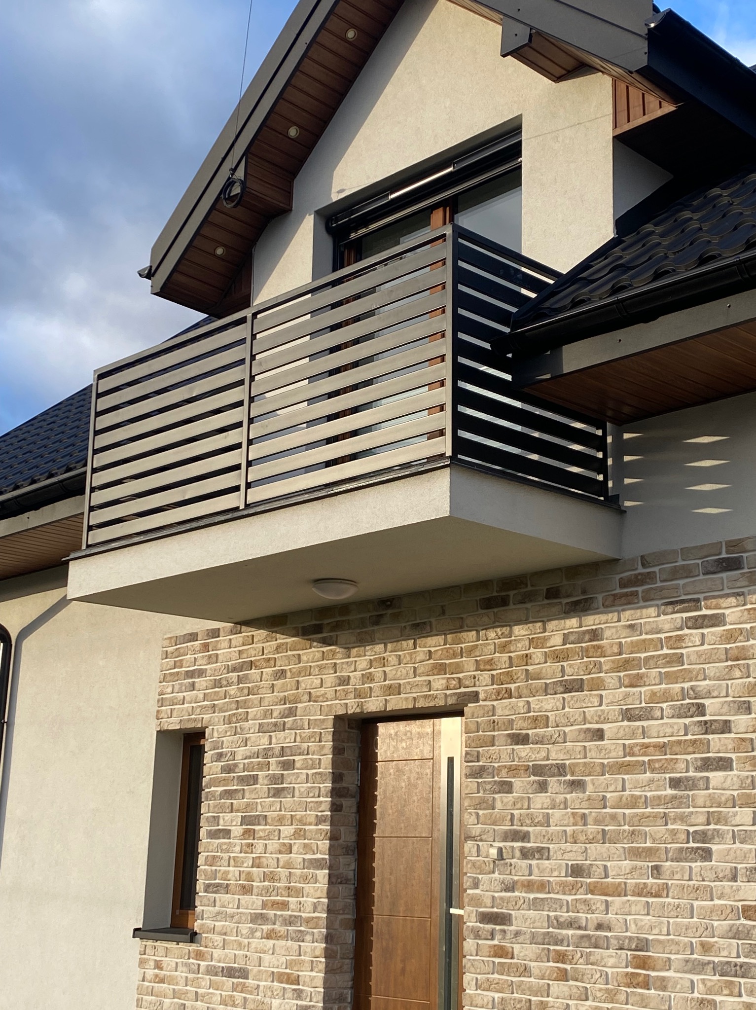 Nowoczesny balkon z poziomą balustradą z metalowych profili na tle elewacji z cegły klinkierowej i jasnego tynku.