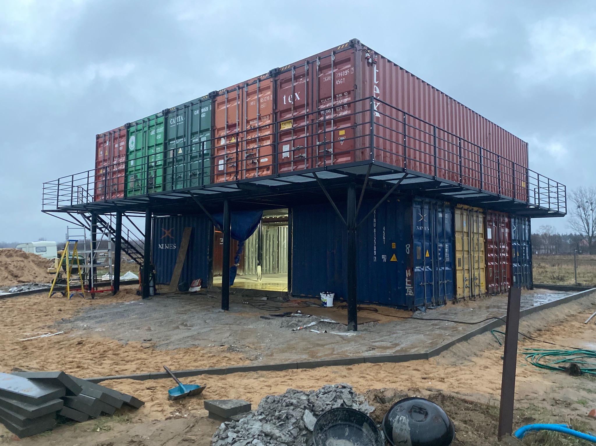 Dwukondygnacyjny budynek z kontenerów morskich w trakcie budowy, z metalową konstrukcją balkonu i schodami, otoczony piaskiem i materiałami budowlanymi.