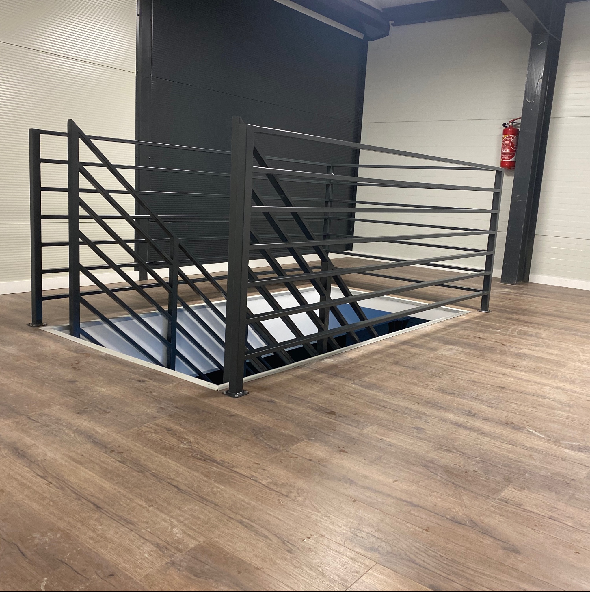 Metalowa barierka zabezpieczająca otwór w podłodze w hali przemysłowej, widoczny gaśnica na ścianie w tle.